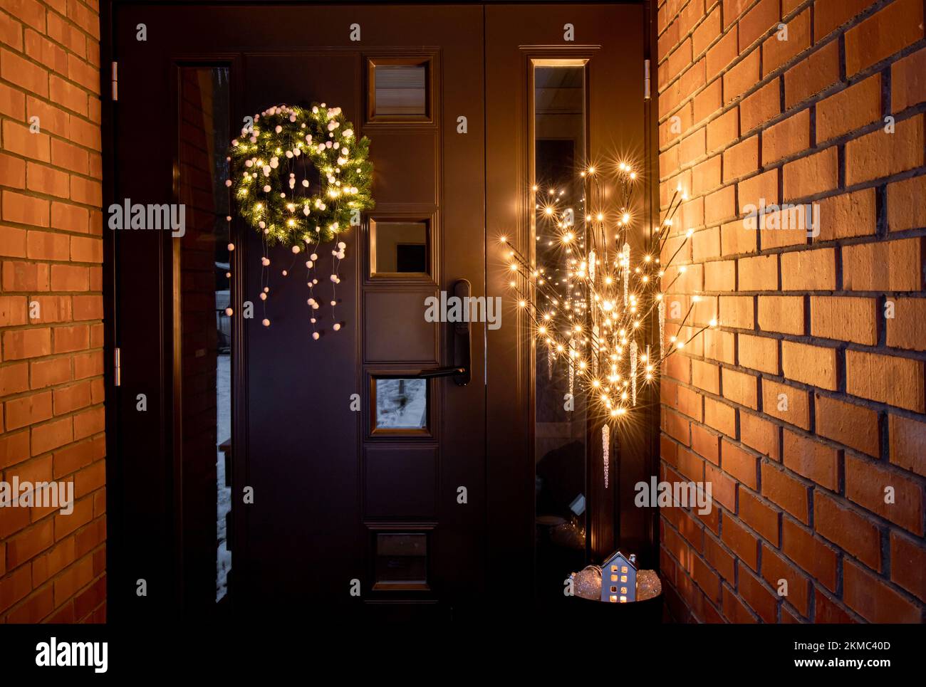 Haustür Außeneingang mit verschiedenen Weihnachtsdekorationen Weihnachtskranz mit Pompons und Blumentopf mit künstlichem Baum mit Lichtern. Stockfoto