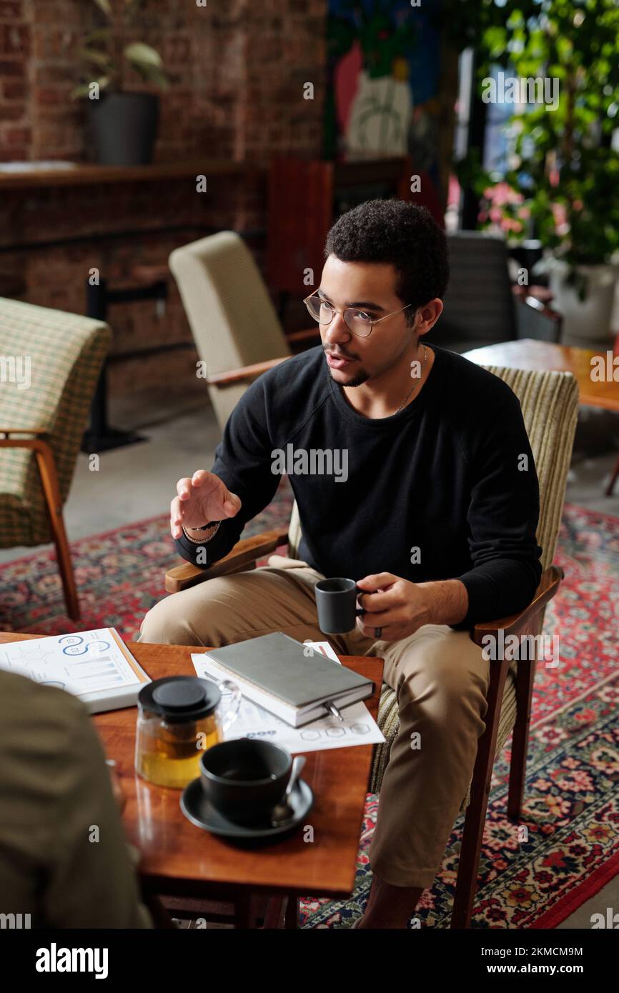 Selbstbewusster Makler in Sachen Freizeitkleidung und Brille, Tee trinken und mit Kollegen kommunizieren, während er in Sesseln für Tische sitzt Stockfoto