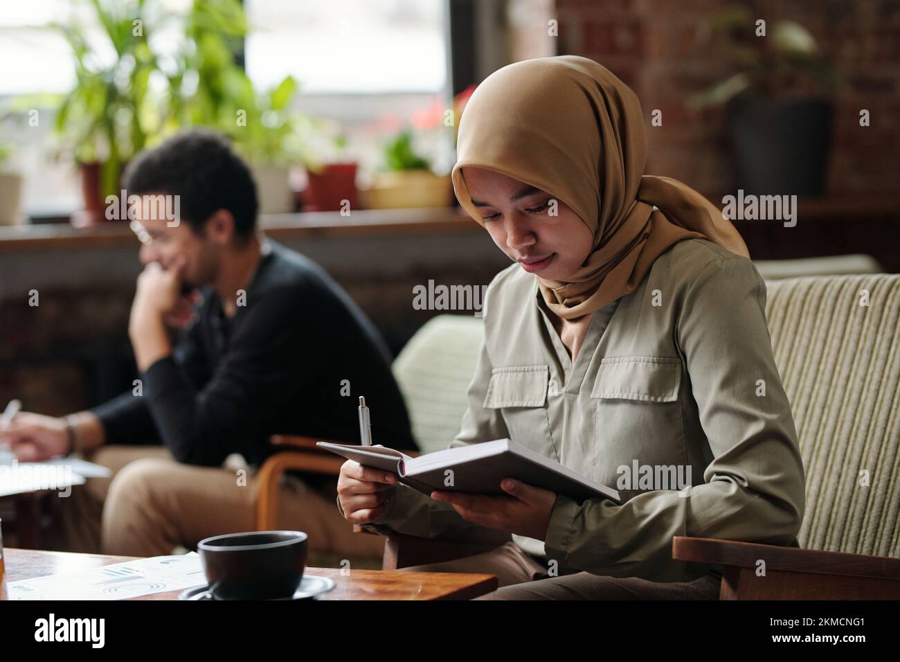 Junge, seriöse muslimische Geschäftsfrau im Hijab, hält ein offenes Notizbuch vor sich und durchsucht Notizen am Arbeitsplatz Stockfoto
