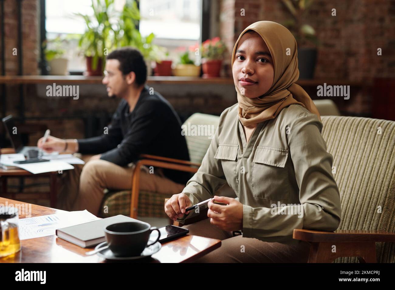 Junge, hübsche Geschäftsfrau im Hijab, sitzt am Tisch vor der Kamera und sieht dich an, während sie Arbeit gegen einen Kollegen organisiert Stockfoto