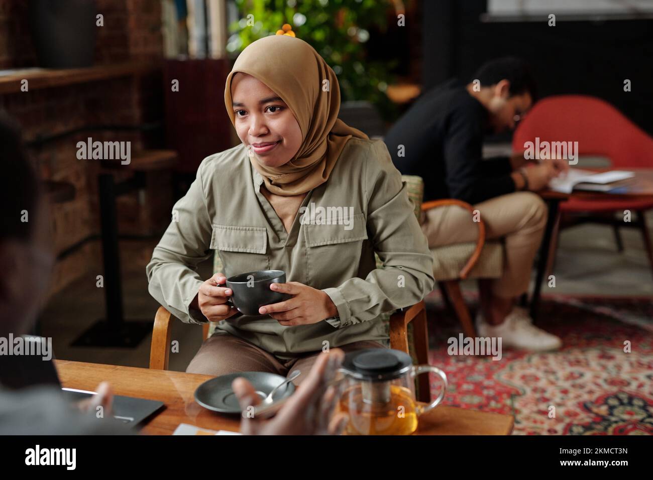 Junge muslimische Mitarbeiterin im Hidschab hält eine Tasse Kräutertee, während sie während des Gesprächs vor einem männlichen Kollegen am Tisch sitzt Stockfoto
