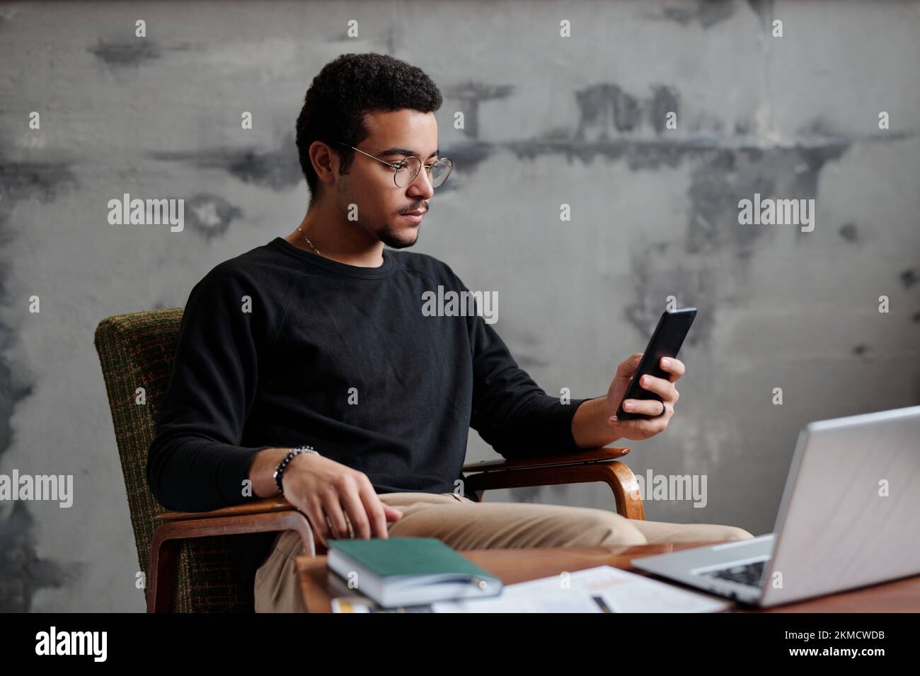 Junger, seriöser Geschäftsmann, der auf den Smartphone-Bildschirm schaut, während er im Sessel am Arbeitsplatz an der grauen Wand im Büro sitzt und SMS schreibt Stockfoto