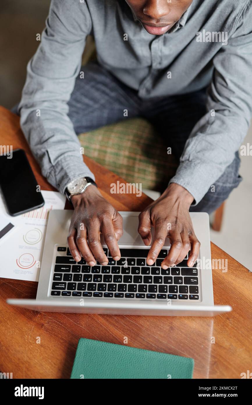 Über dem Blickwinkel eines jungen afroamerikanischen Ökonomen oder Analysten, der die Laptop-Tastatur in die Hand nimmt, während er am Schreibtisch sitzt und seine Arbeit organisiert Stockfoto