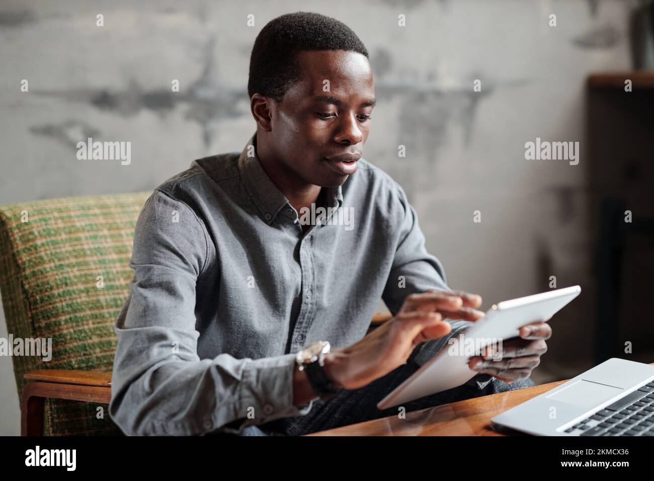 Junger seriöser afroamerikanischer Geschäftsmann in eleganter Freizeitkleidung, der ein Tablet benutzt, während er am Arbeitsplatz im Sessel sitzt und die Arbeit organisiert Stockfoto