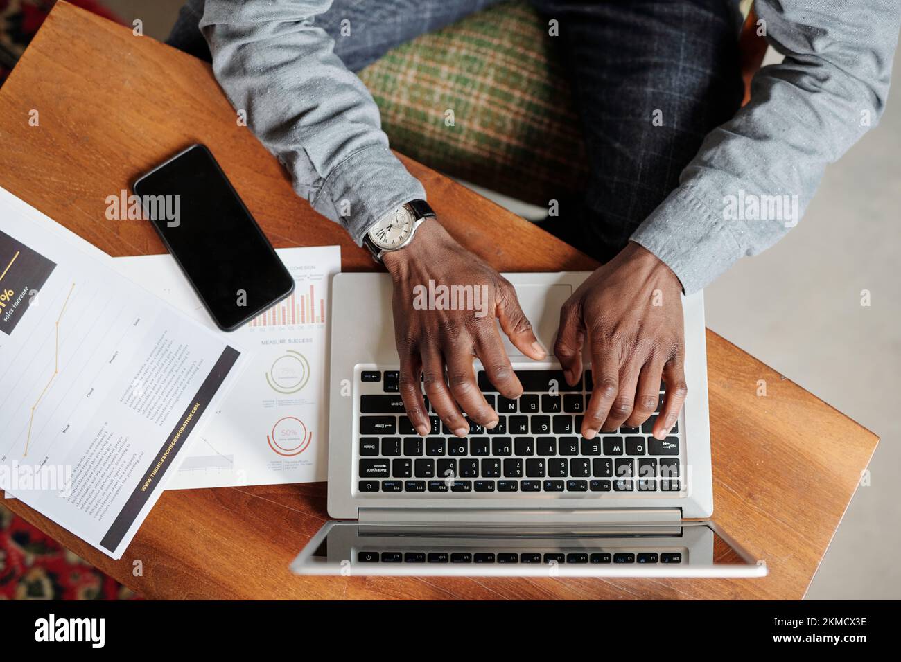Draufsicht der Hände eines jungen Schwarzen, der die Tasten der Laptop-Tastatur drückt, während er mit Smartphone und Finanzunterlagen am Schreibtisch sitzt Stockfoto