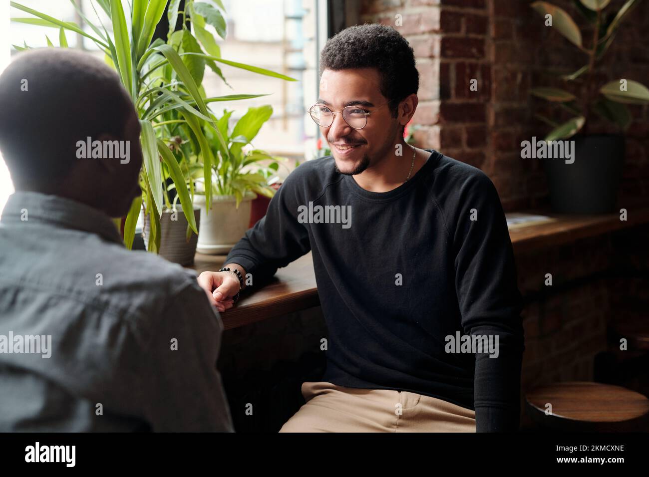 Ein junger lächelnder Geschäftsmann in Freizeitkleidung, der einen afroamerikanischen männlichen Kollegen mit einem Lächeln ansieht, während er über Organisationspunkte spricht Stockfoto