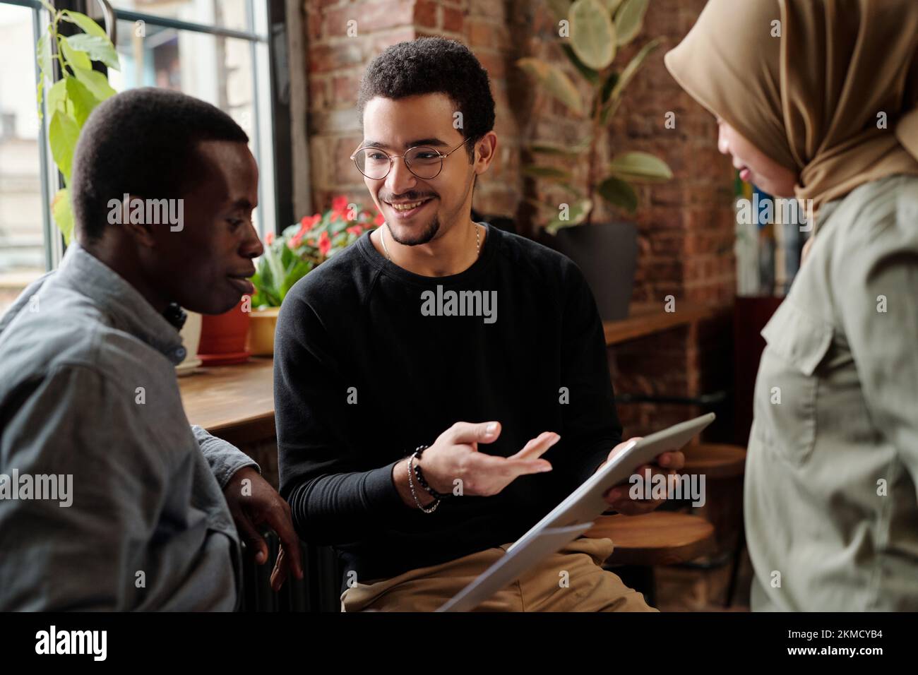 Selbstbewusster junger Mitarbeiter mit Tablet, der interkulturellen Kollegen während des Arbeitsgesprächs ein neues Unternehmensprojekt vorstellt Stockfoto