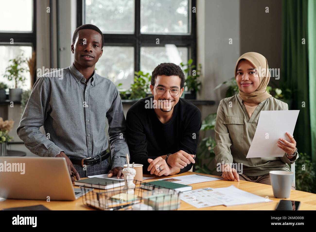 Gruppe von drei glücklichen jungen interkulturellen Mitarbeitern in Freizeitkleidung, die mit Dokumenten und Laptop vor die Kamera schauen, während sie am Arbeitsplatz stehen Stockfoto