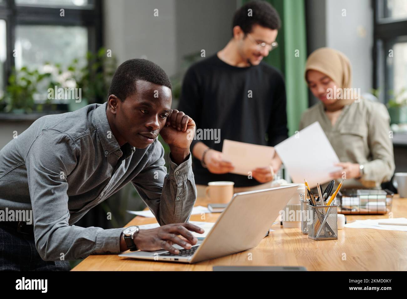 Junger afroamerikanischer Makler, der sich vor dem Laptop über den Arbeitsplatz beugt, während er Online-Daten analysiert oder Arbeit gegen Kollegen organisiert Stockfoto