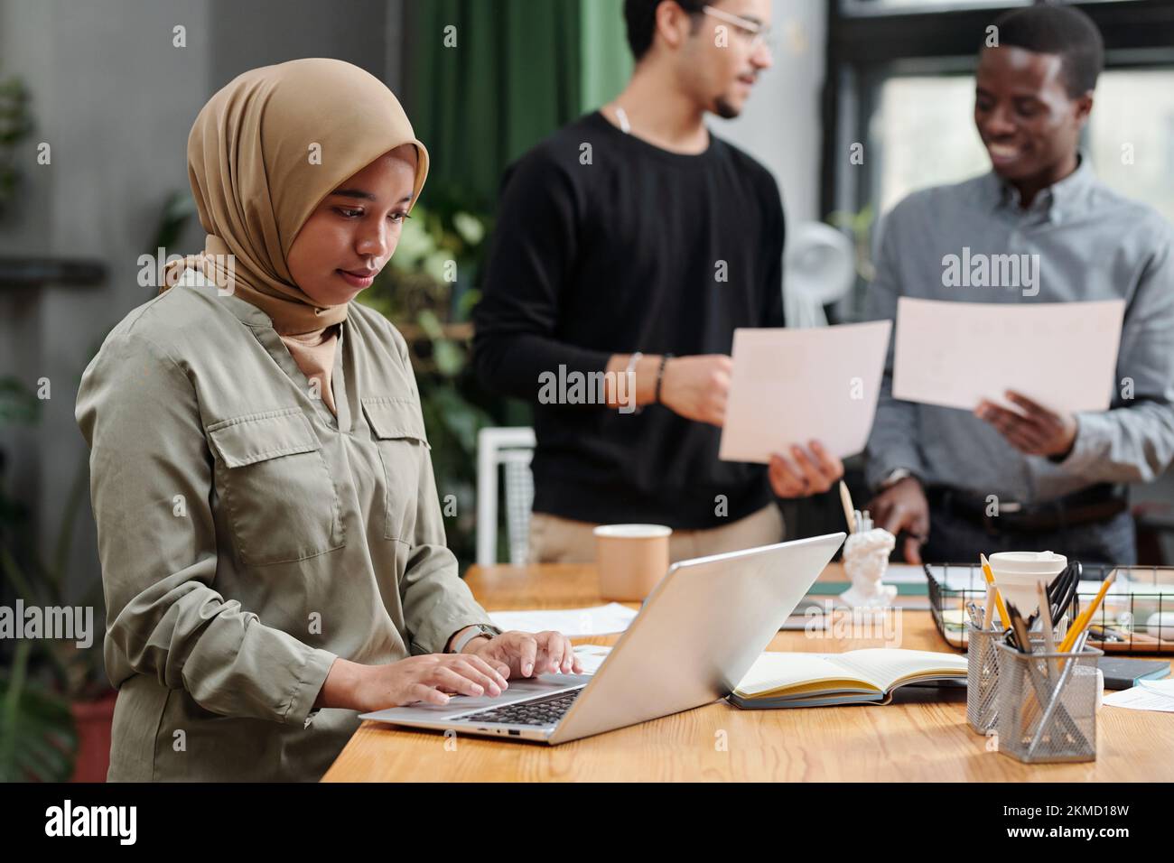 Junge muslimische Geschäftsfrau, die ihre Arbeit vor dem Laptop organisiert, während sie am Arbeitsplatz gegen zwei interkulturelle Männer mit Papieren steht Stockfoto