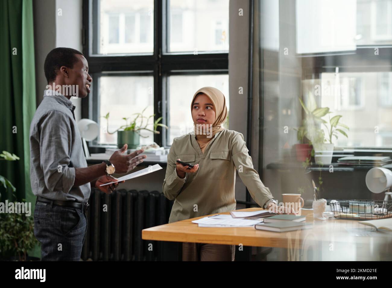 Junge muslimische Geschäftsfrau, die zuversichtlichem afroamerikanischen männlichen Kollegen zuhört, der bei der Sitzung einen Bericht über die Erstellung von Dokumenten vorlegt Stockfoto