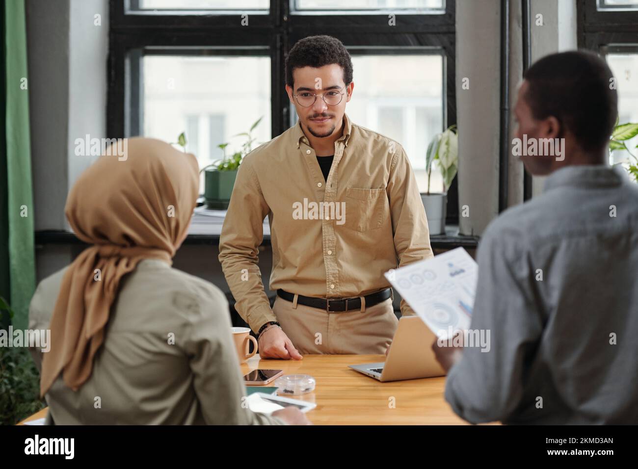 Junge, ernste Managerin, die sich Berichte über eine muslimische Untergebene anhört, während sie bei der Besprechung vor zwei interkulturellen Mitarbeitern steht Stockfoto