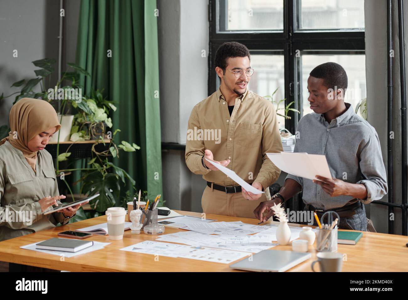 Zwei junge interkulturelle Geschäftsleute diskutieren über Finanzpapiere, während ihre Kollegin bei einem Arbeitstreffen im Büro ein Tablet benutzt Stockfoto