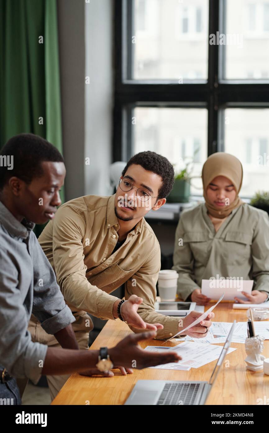 Junger, zuversichtlicher männlicher Ökonom mit Finanzdokument, der afroamerikanische Kollegen während der Diskussion grafischer Daten ansieht Stockfoto