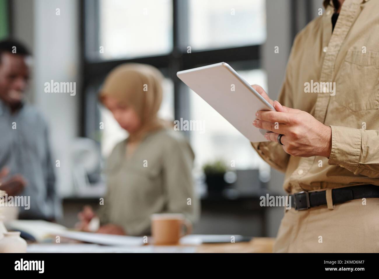 Hände eines jungen Analysten oder Maklers, der ein digitales Tablet verwendet, während er vor der Kamera steht und Kollegen über Arbeitspläne diskutieren Stockfoto