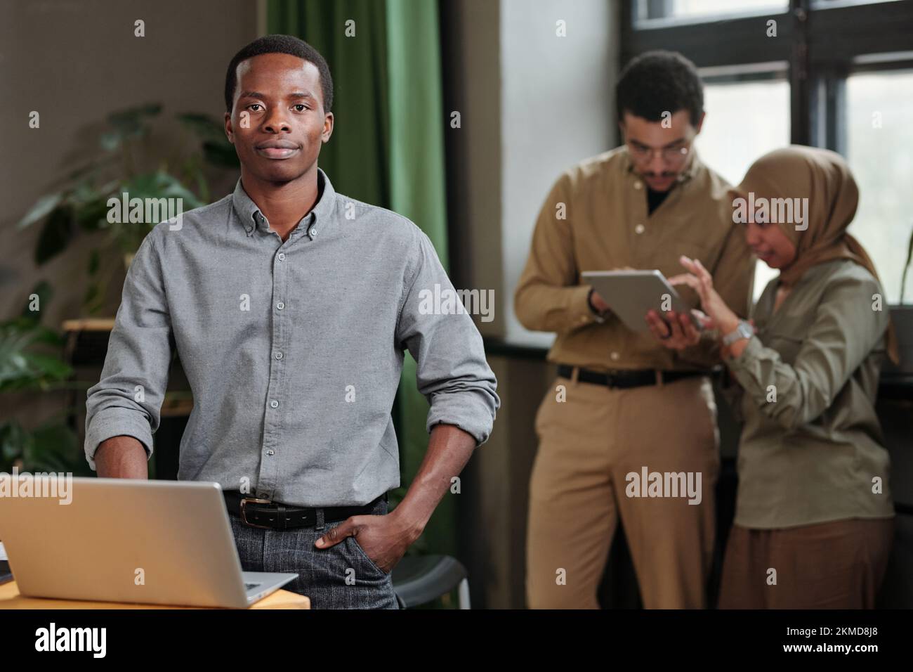 Junger, selbstbewusster Geschäftsführer in intelligenter Freizeitkleidung, der am Arbeitsplatz vor der Kamera gegen zwei Kollegen steht Stockfoto