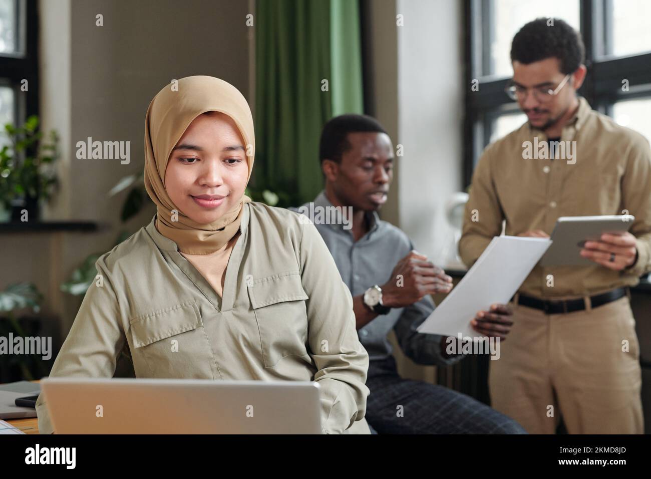 Junge muslimische Geschäftsfrau in Hidschab und Freizeitkleidung, die vor einem Laptop sitzt und sich mit zwei interkulturellen männlichen Mitarbeitern vernetzt Stockfoto