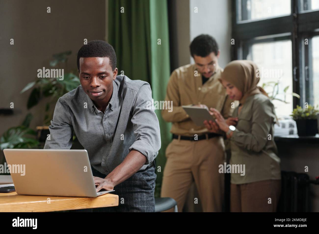 Junger afroamerikanischer Geschäftsmann, der sich mit einem Notebook über den Schreibtisch beugt und sich mit zwei interkulturellen Kollegen über ein Tablet vernetzt Stockfoto