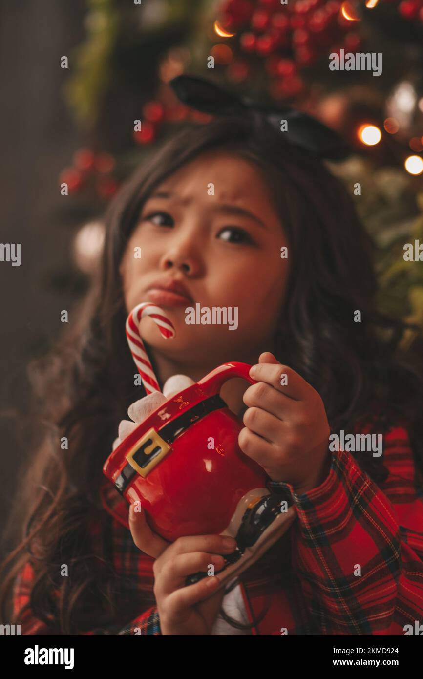 Happy japan kleines Mädchen Träumer warten Wunder Santa genießen Süßigkeiten in der Nähe von noel Baum. Asiatische Kind in rot kariert Outfit feiert Neujahr halten Xmas Cup wi Stockfoto