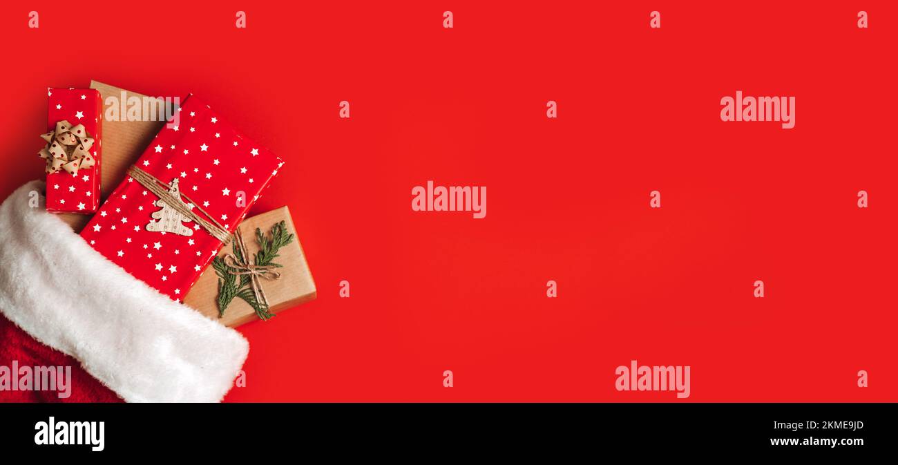Weihnachtsmann-Hut mit Geschenkboxen auf rotem Hintergrund. Weihnachtsgeschenke und weihnachtsmannmütze auf rotem Hintergrund. Geschenkideen für Weihnachten Stockfoto