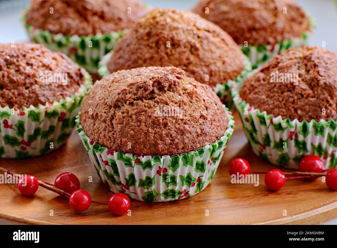 Heiße Bran-Muffins in Holly Backbechern für den Weihnachtsmorgen. Auf Holzplatte mit roten Beeren. Selektiver Fokus auf Muffin in der Mitte vorn. Stockfoto