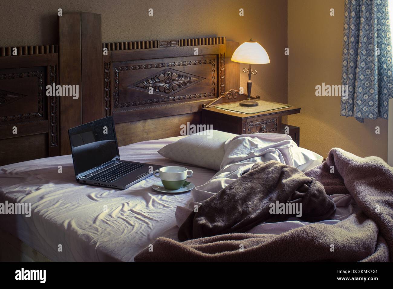 Ein rustikales Schlafzimmer mit einer Tasse Suppe auf einem schmutzigen Teller neben einem offenen Laptop auf einem ungemachten Bett mit Decken, die von einer Nachttischlampe beleuchtet werden und Sonnenlicht vom Stockfoto
