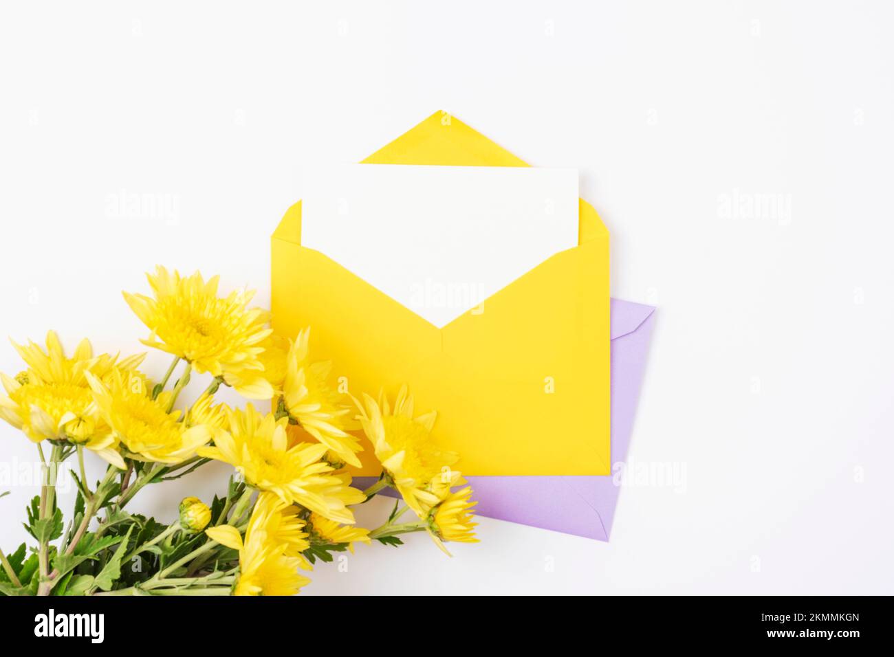 Gelber Umschlag mit leerer Karte und Blumen auf weißem Hintergrund. Geburtstag, Einladung, Feiertagskonzept. Draufsicht, flaches Lay, Modell. Stockfoto