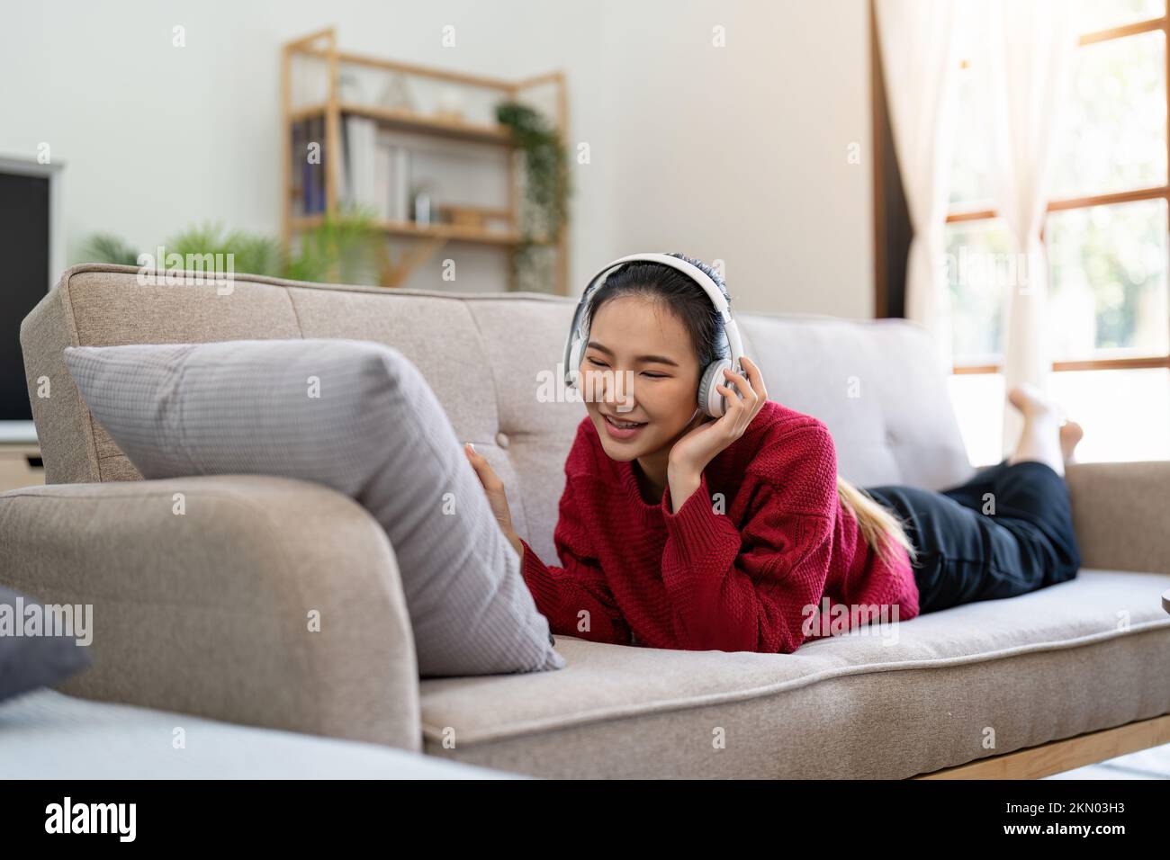 Eine sorglose Asiatin, die Musik hört, Kopfhörer benutzt, auf dem Sofa im Wohnzimmer liegt, einen Kopierraum. Glückliche junge Dame mit geschlossenen Augen, die Spaß hat Stockfoto
