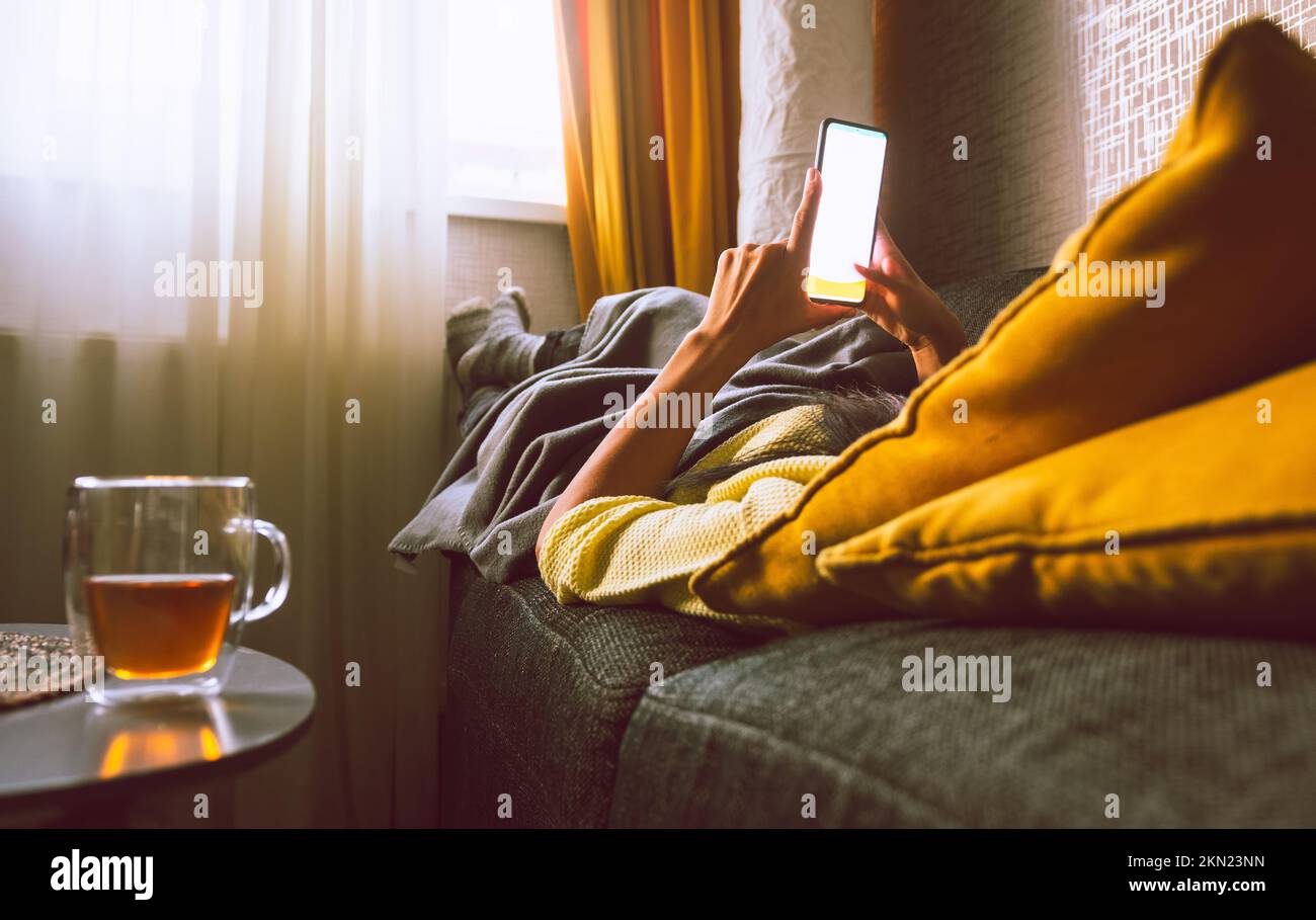 Die Person liegt im Bus mit dem Smartphone mit leerem Kopierbildschirm. Genießen Sie im Winter Tee mit den Füßen auf der Heizung im kalten Haus. Zentralheizung und en Stockfoto