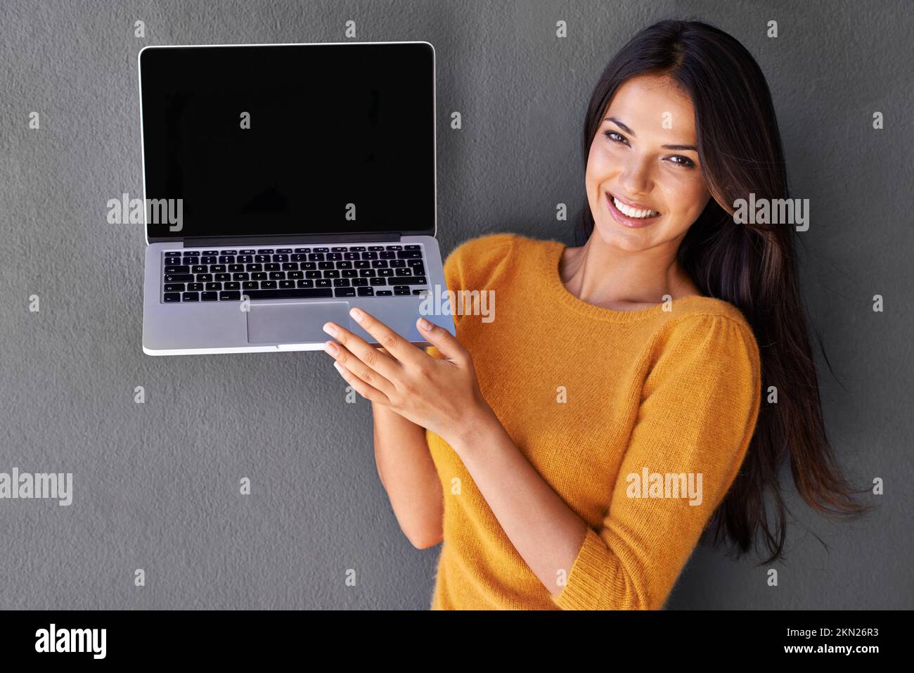 Sie ist ein sozialer Online-Schmetterling. Eine schöne Frau, die einen leeren Laptop-Bildschirm vor grauem Hintergrund präsentiert. Stockfoto