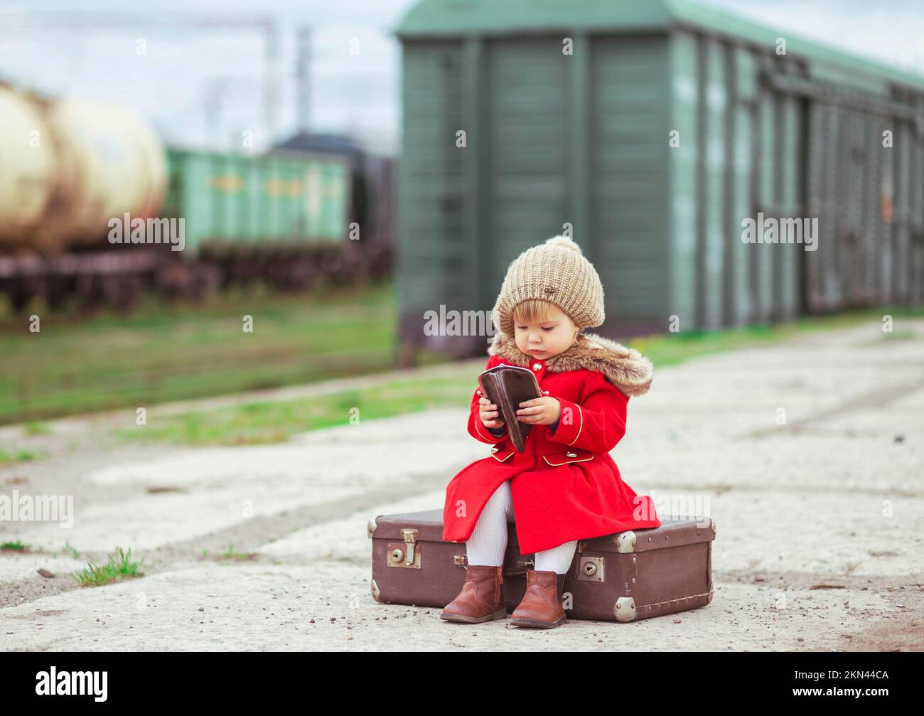 Ein bezauberndes Baby in einem roten Mantel mit Koffer wartet auf einen Zug Stockfoto