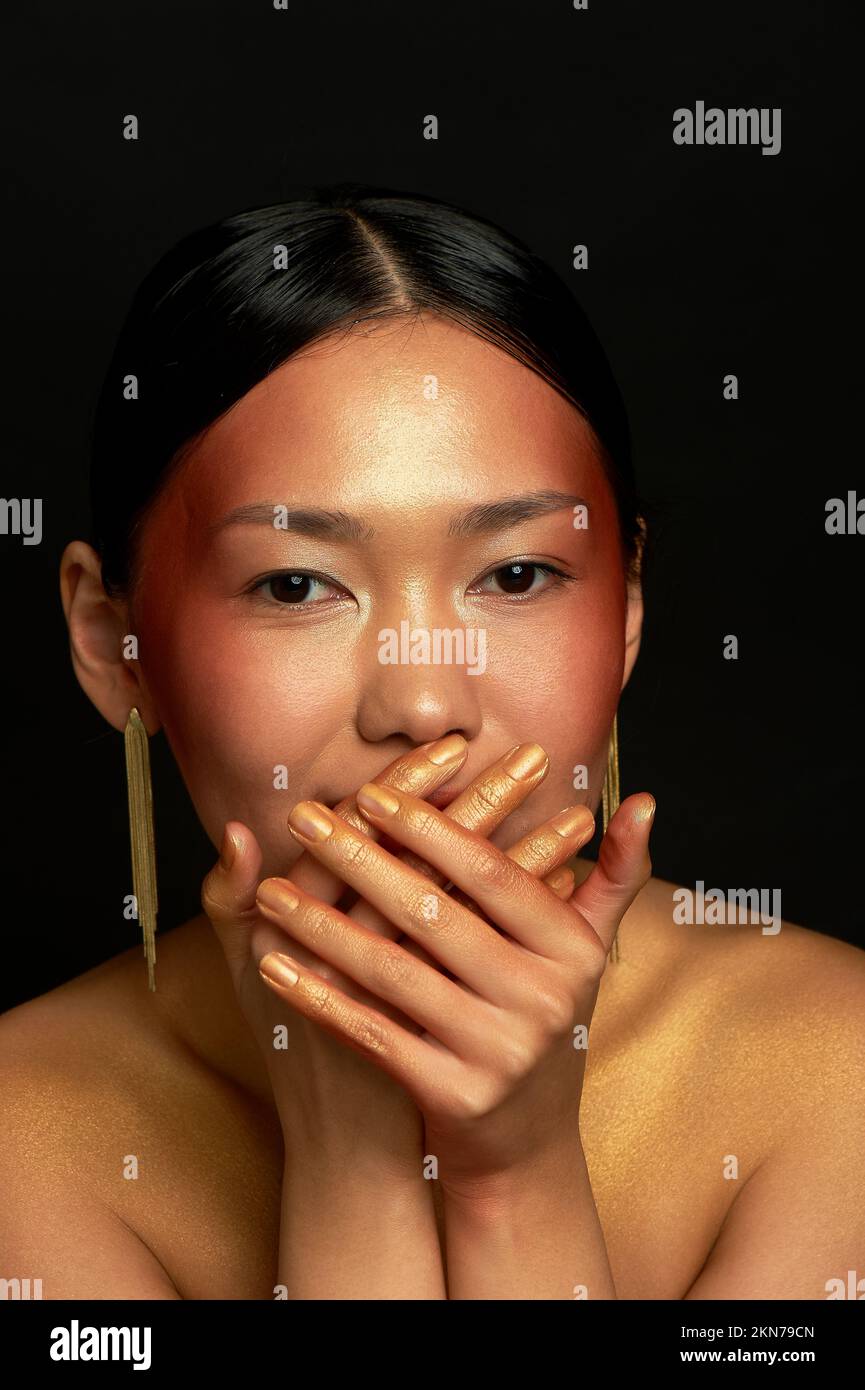 Ein asiatisches Mädchen mit schwarzem Hintergrund im Studio. Gold. Verbirgt ein Lächeln Stockfoto