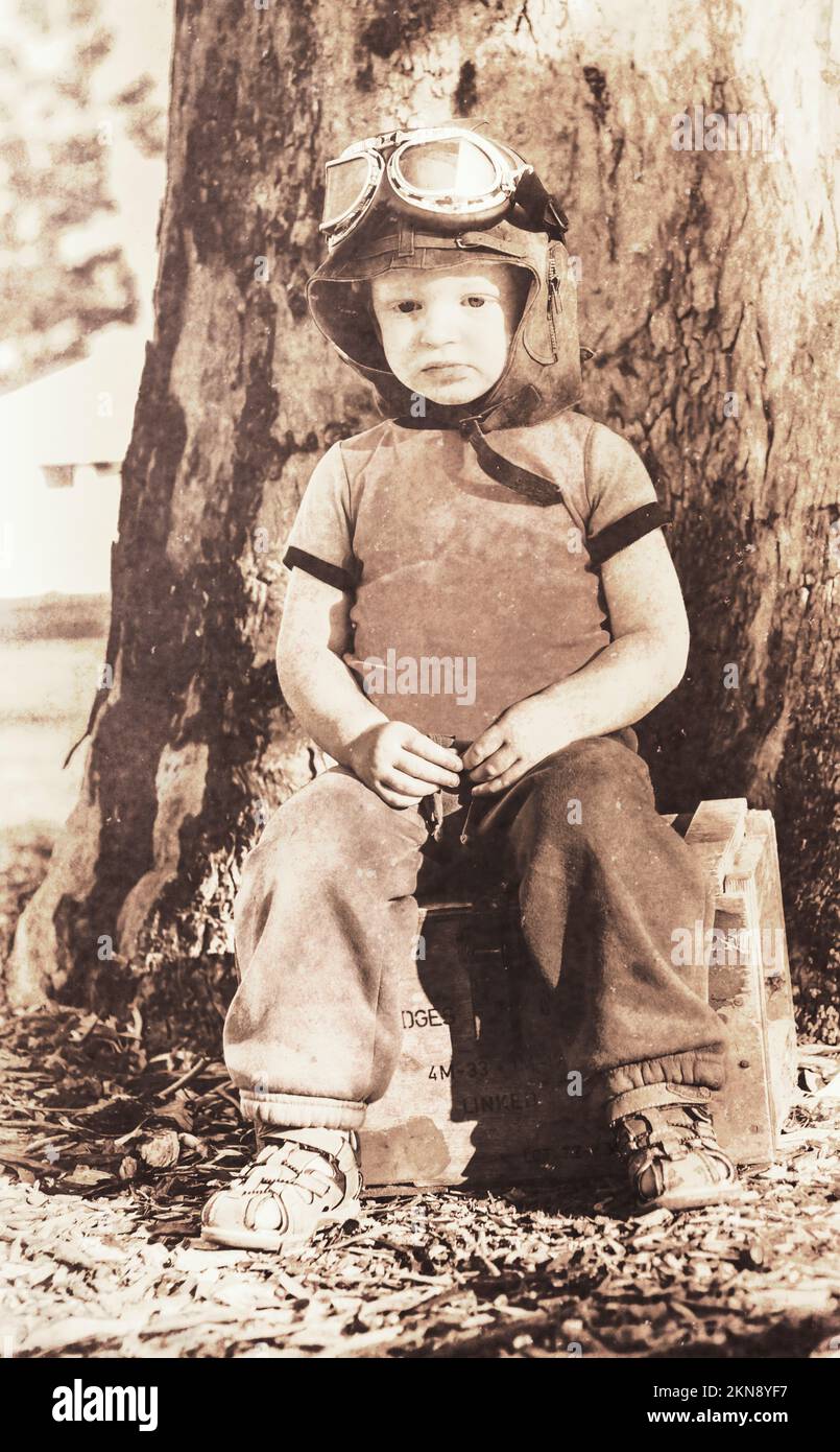 Vintage 1940 Stil Bild von einem niedlichen 3yo Kind trägt Flugbrille, während auf einer Munitionskiste sitzen. ww2 Flügelmann hält die Forte Stockfoto
