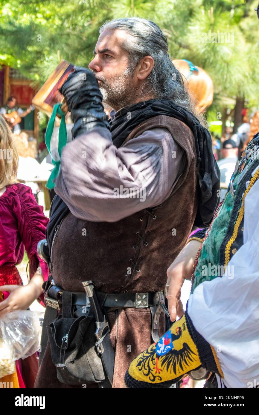 Ein Mann in Kostümen mit langen grauen Haaren trinkt zusammen mit anderen Fair-Stars im Hintergrund beim Oklahoma Renaissance Festival Muskogee Okla einen Drink aus einem Holzbecher Stockfoto