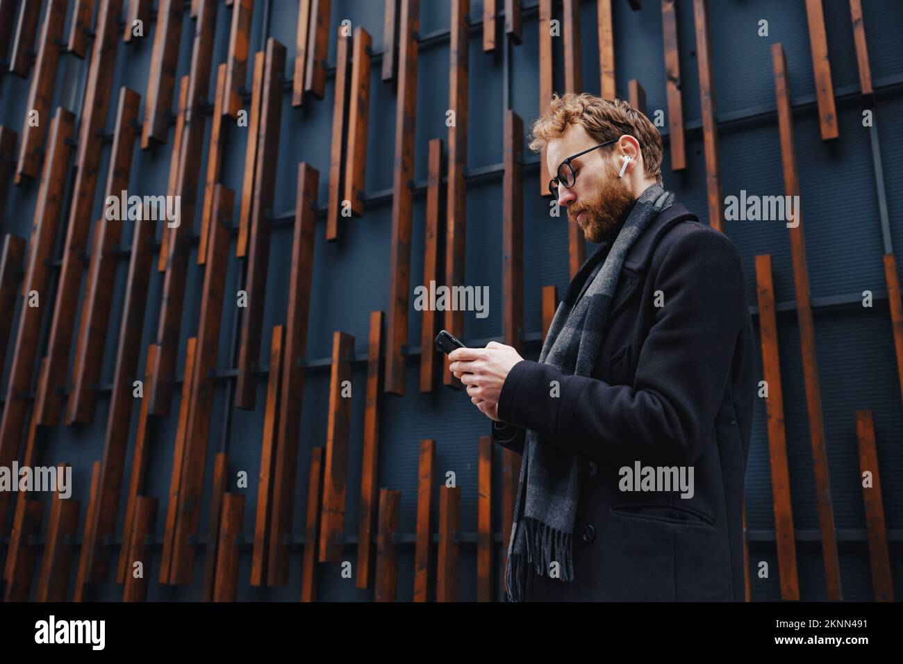 Ein junger rothaariger Typ, ein Geschäftsmann mit Brille, vor dem Hintergrund des urbanen Stils, überprüft Post auf einem Smartphone, kommuniziert in einem Boten und Stockfoto