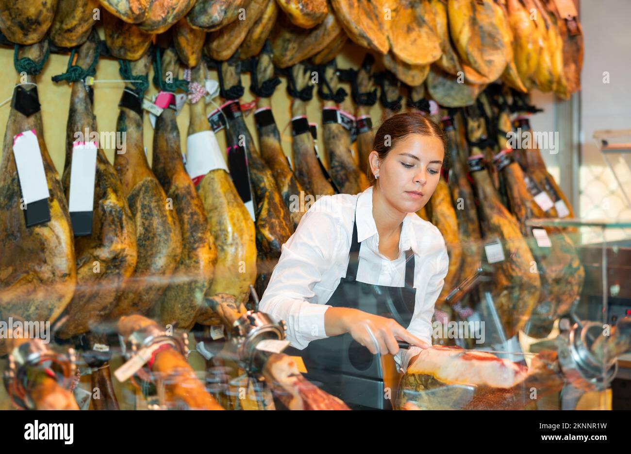 Junge Verkäuferin in schwarzer Schürze schneidet iberischen Pyjon an der Theke in der Metzgerei Stockfoto