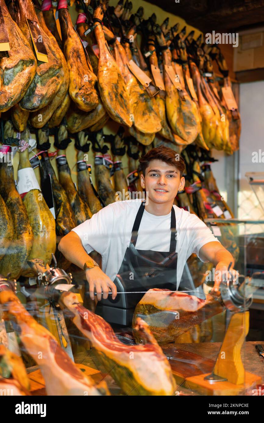 Junger Verkäufer in einer Schürze, der iberischen Jamon auf jamonera in der Metzgerei zerschneidet Stockfoto