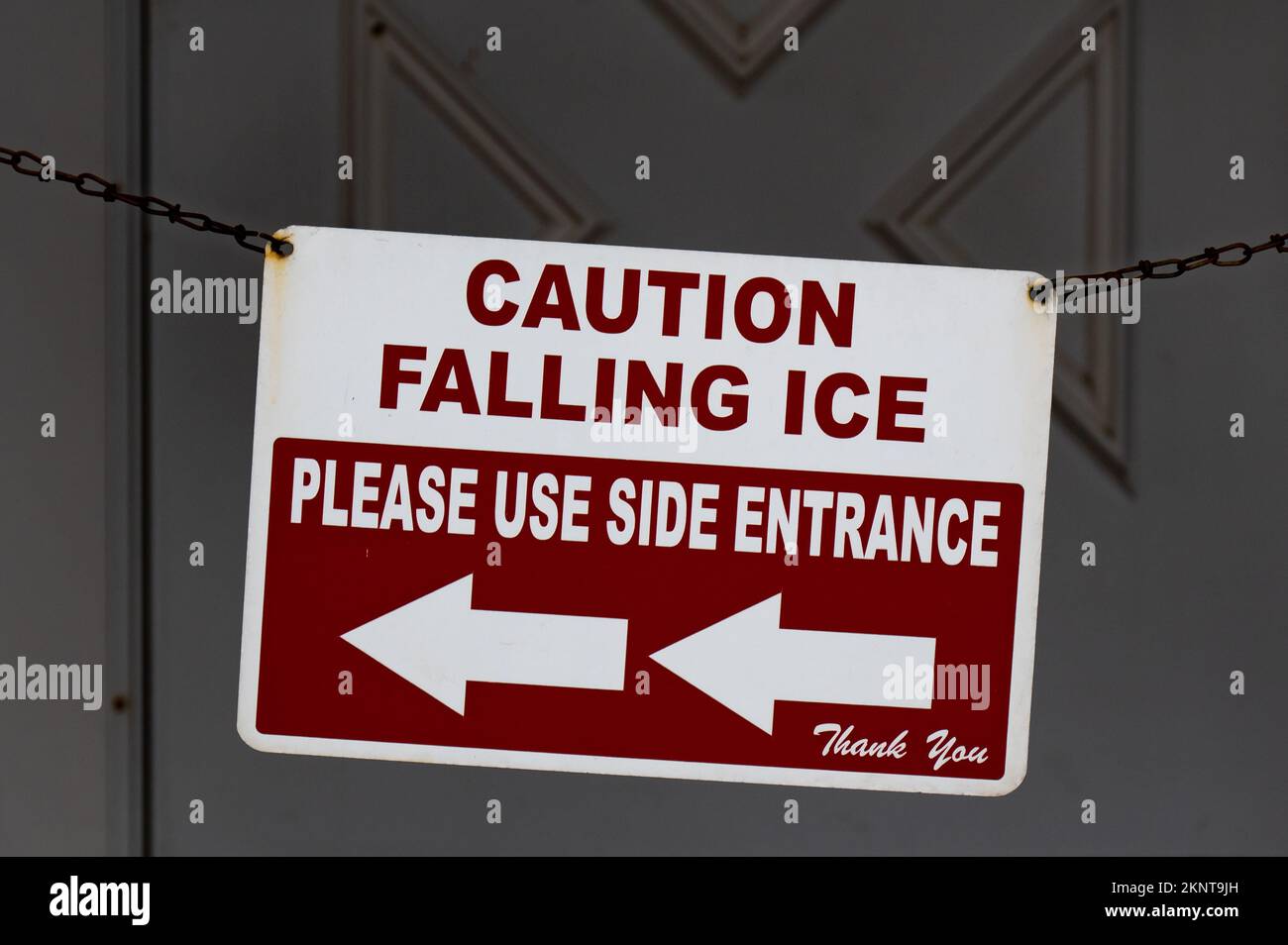 Ein rot-weißes Schild mit der Aufschrift „Vorsicht: Fallendes Eis“ an einer Kette auf der Veranda eines Gebäudes, das den Verkehr zum Seiteneingang leitet. Stockfoto