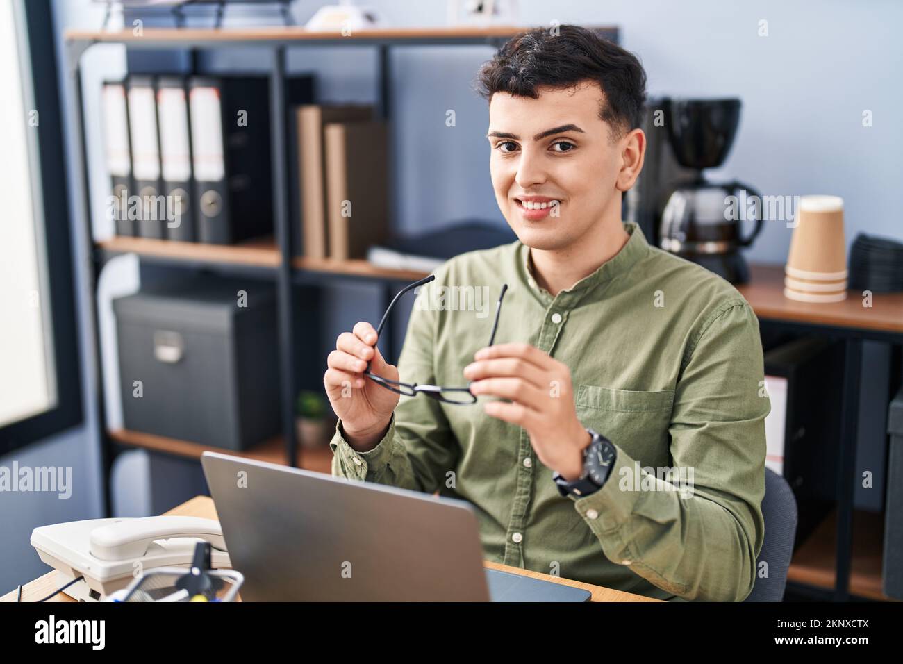 Junger nicht-binärer Mann, der im Büro eine Brille mit einem Laptop hält Stockfoto