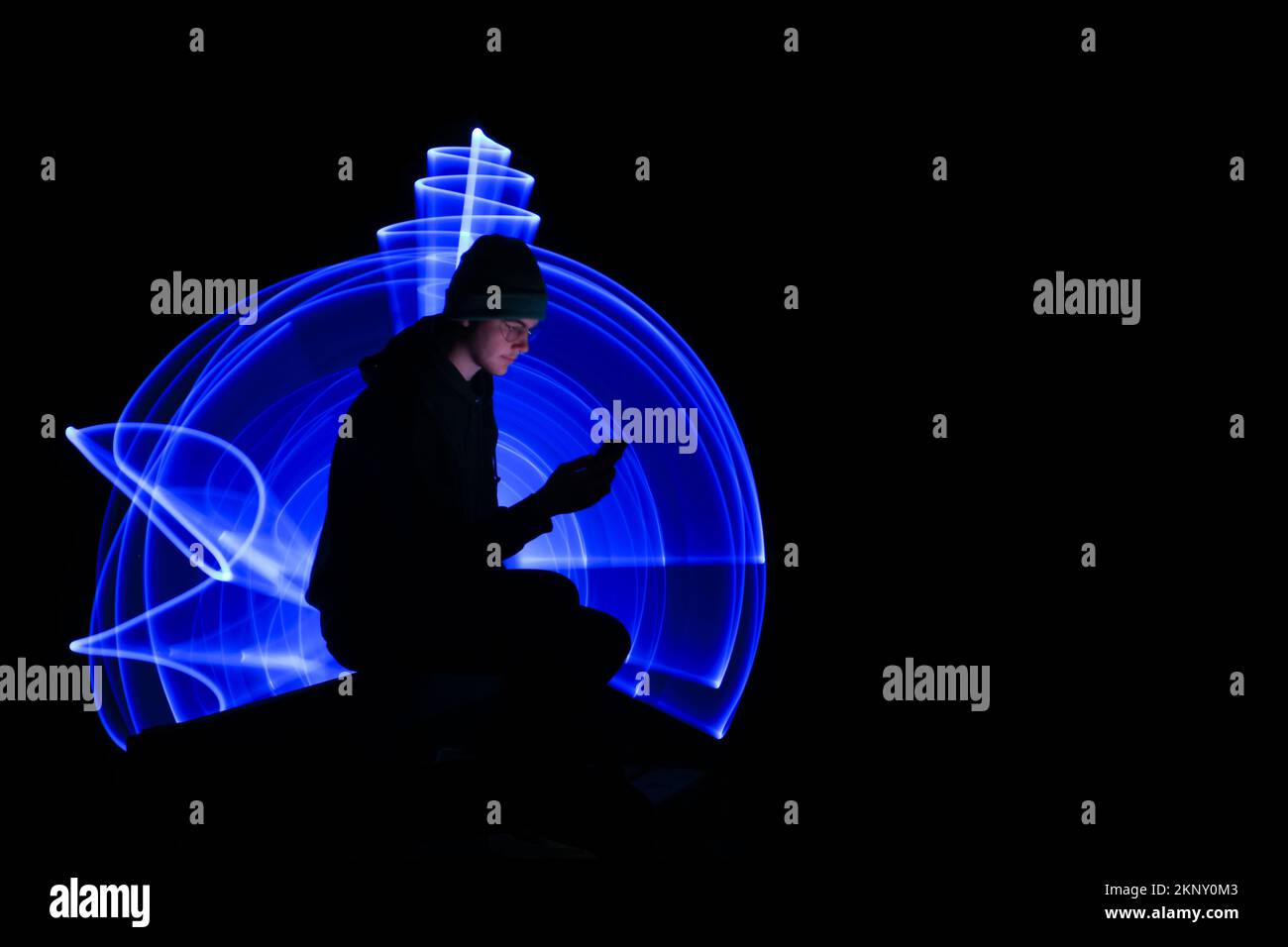 Ein junger Mann, der auf einer Bank sitzt und sein Handy ansieht und benutzt. Szene im Hintergrund mit blauem Licht beleuchten. Eine Person mit Smartphone. Stockfoto