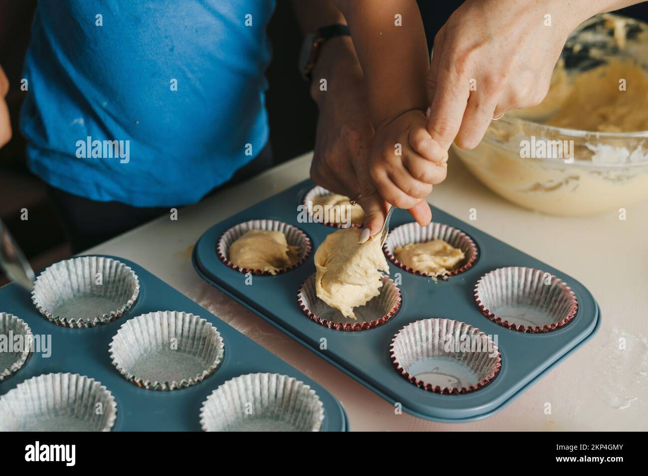 Die Hände einer Mutter und eines Säuglings lernen, wie man Muffindosen mit Teig mit einem Löffel füllt. Süßes Essen. Familienkochen zusammen. Stockfoto