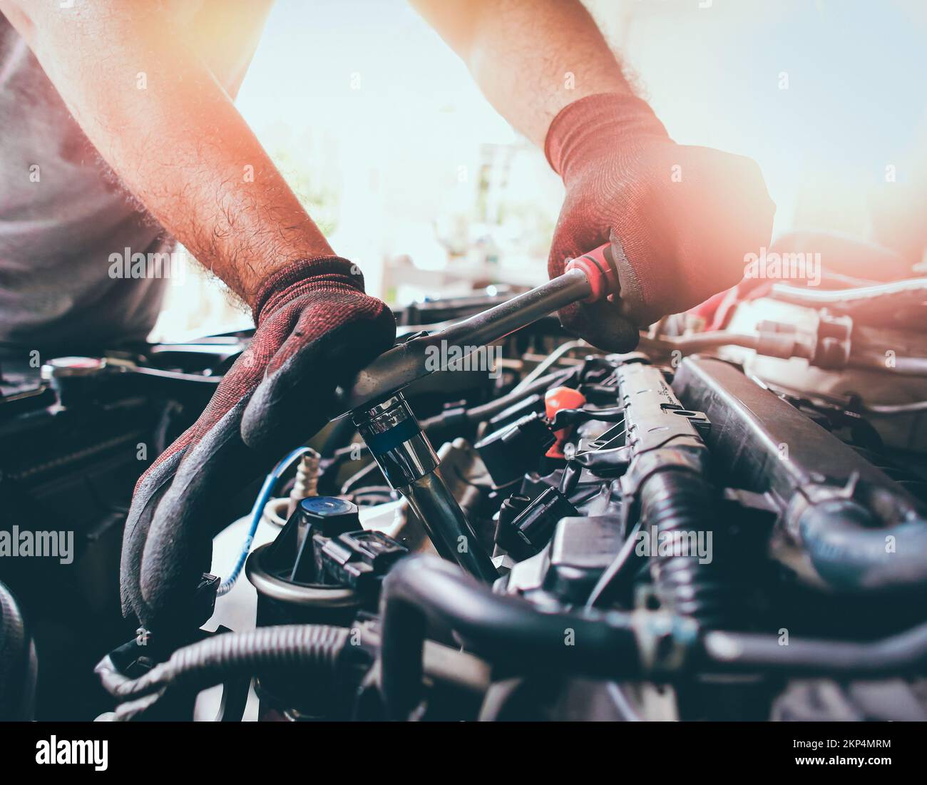 Die rote Handschellen des Automechanikers befestigen den Bolzen mit dem Steckschlüssel, um den Fahrzeugmotor zu fixieren, Sonnenlicht auf dem Hintergrund Stockfoto