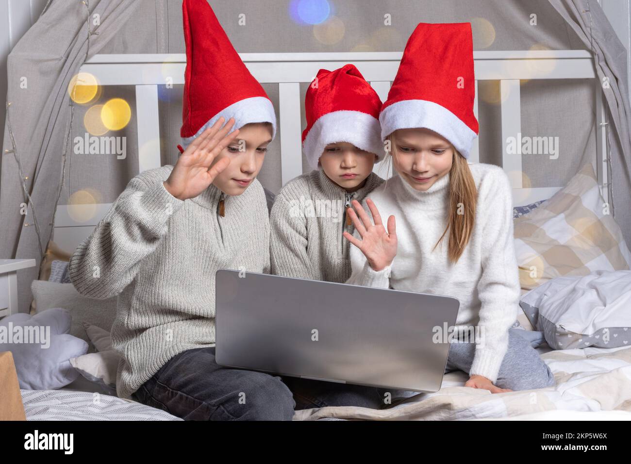 Fröhliche Kinder in roten Weihnachtsmannhüten mit einem Laptop an Heiligabend zu Hause. Jungs und ein Mädchen gratulieren online Verwandten, die auf dem Bett sitzen. Familie x-Mas ev Stockfoto