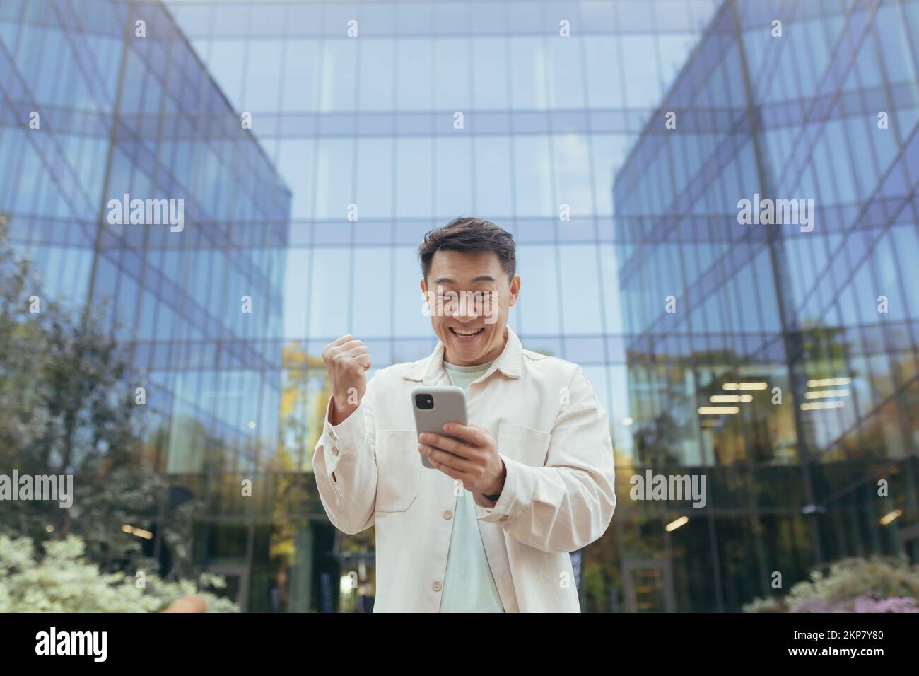 Asiatischer Geschäftsmann in lässiger Kleidung mit Telefon außerhalb des Bürogebäudes, der Sieg und Erfolg feiert, Mann, der fröhlich Hand hoch Triumph Geste hält. Stockfoto