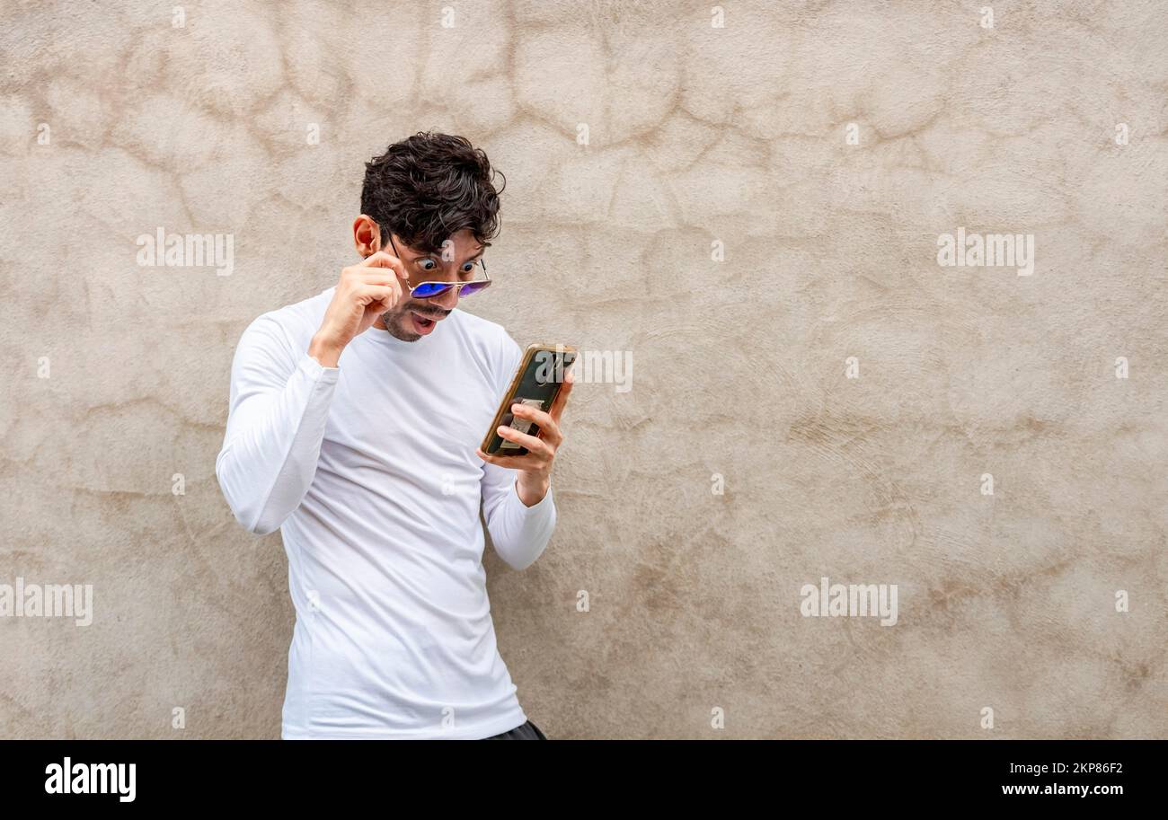 Ein überraschter junger Lateiner, der sein Handy an eine Wand lehnt. Ein hübscher Typ mit Brille, der erstaunt war, wie sein Handy an eine Wand lehnte, Lat Stockfoto
