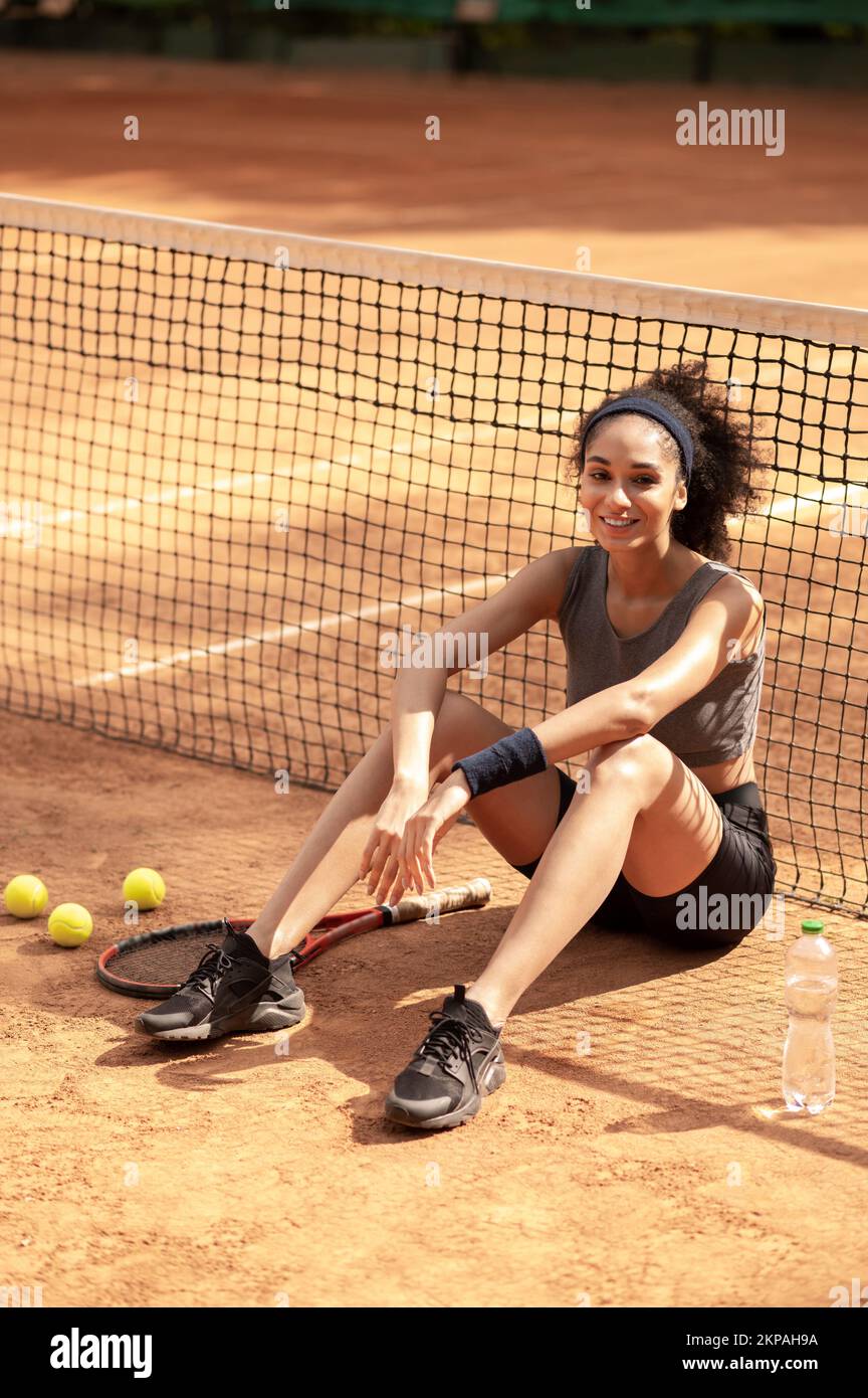 Süßes dunkelhaariges Mädchen, das auf dem Tennisplatz sitzt und zufrieden aussieht Stockfoto