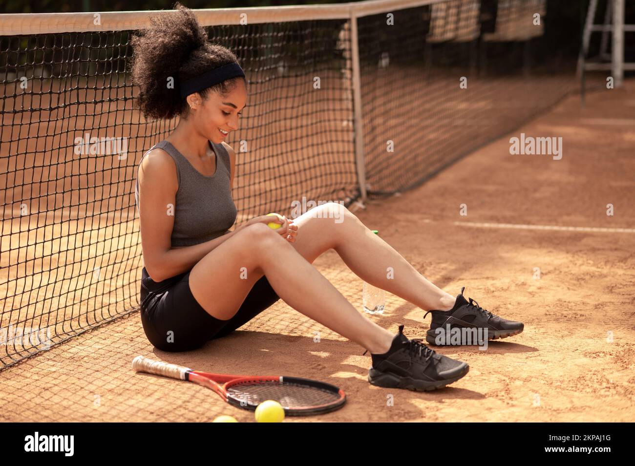 Süßes dunkelhaariges Mädchen, das auf dem Tennisplatz sitzt und zufrieden aussieht Stockfoto