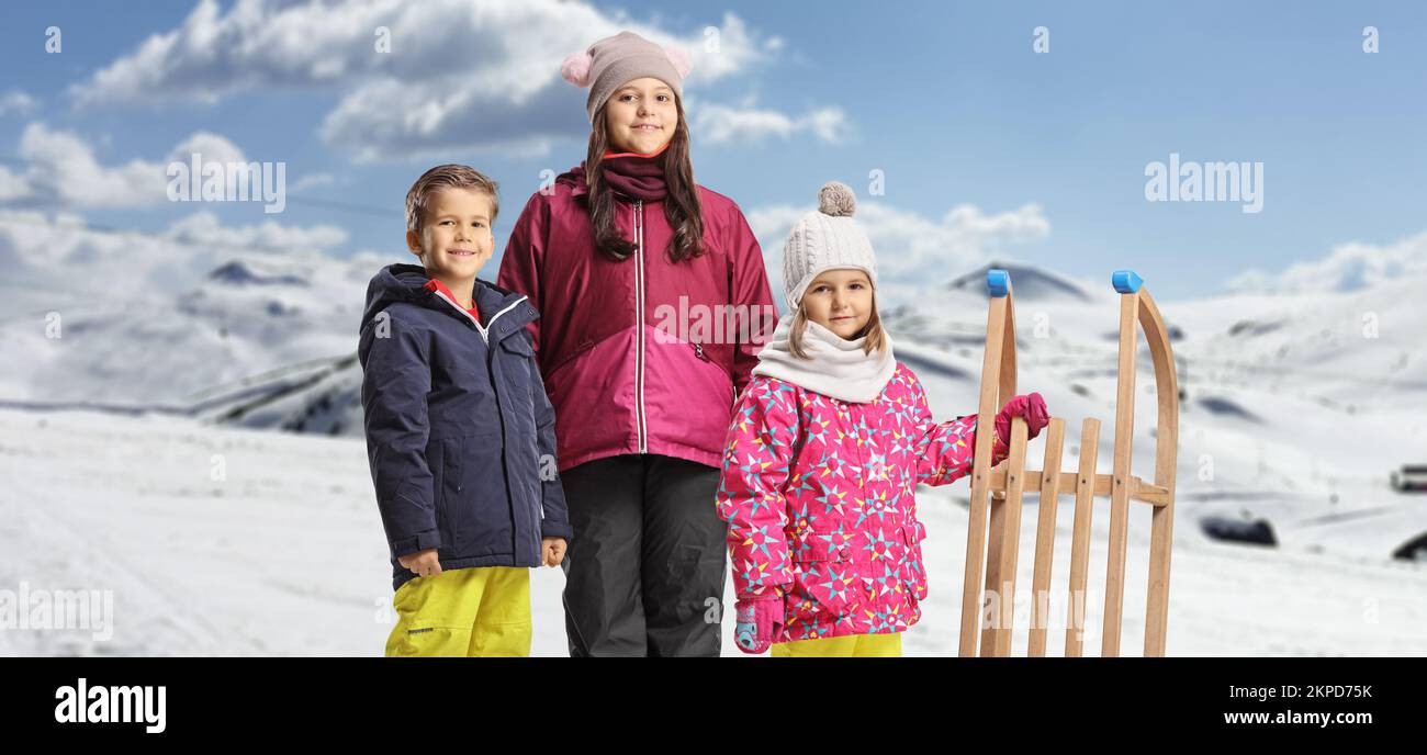 Kinder in Winterkleidung stehen mit einem hölzernen Schlitten auf einem verschneiten Berg Stockfoto