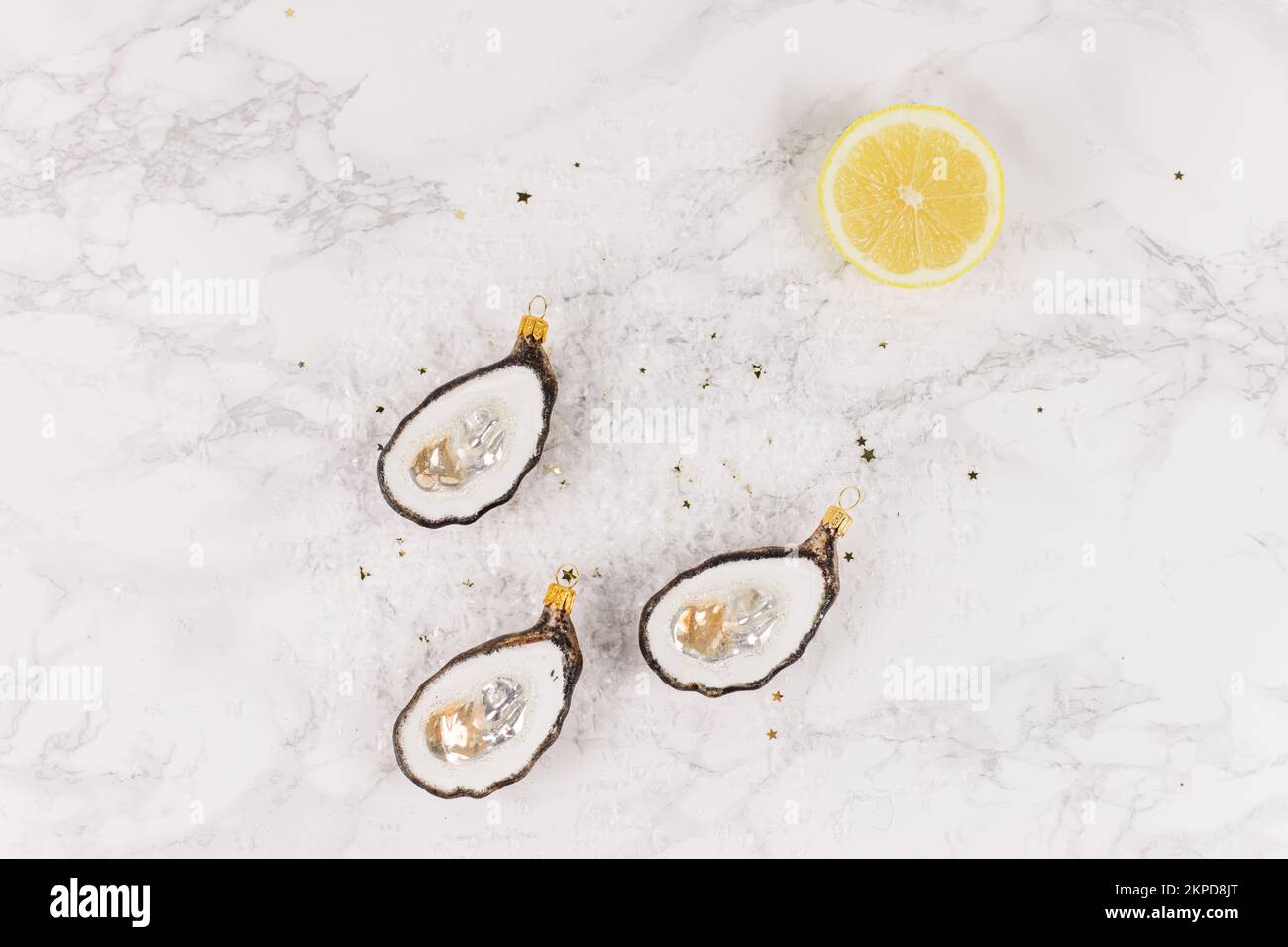 Drei Weihnachtsbaumkugeln in Form einer Auster liegen auf einem Marmortisch. Glitzer, Sterne und Zitronenscheiben schmücken das Bild. Stockfoto