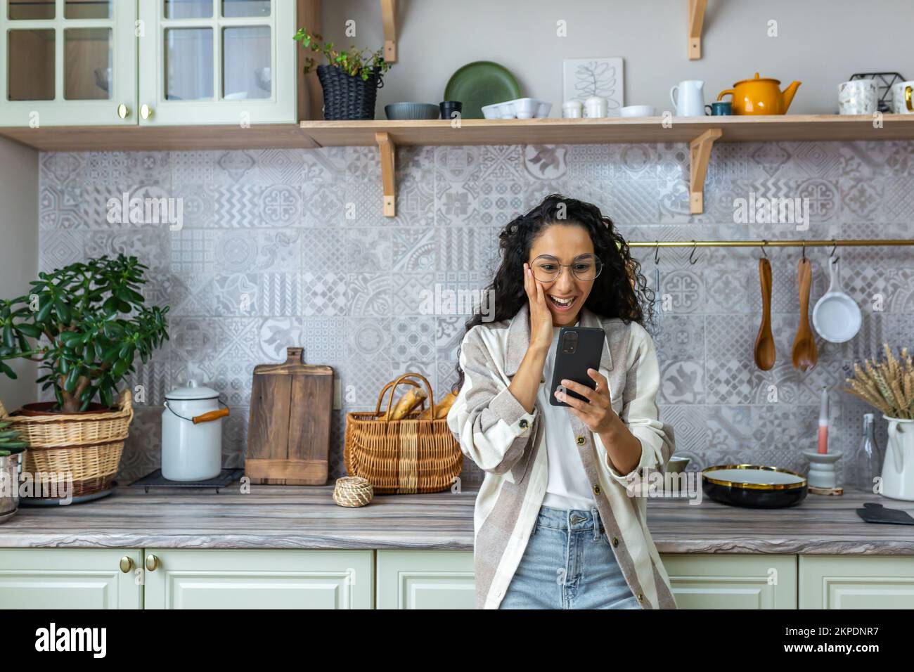 Eine glückliche lateinamerikanische Frau steht zu Hause in der Küche, hält das Telefon in der einen Hand und ihren Kopf mit der anderen Hand. Ich bin froh, dass ich eine unerwartete und freudige Nachricht erhalten habe, eine gute Nachricht. Stockfoto